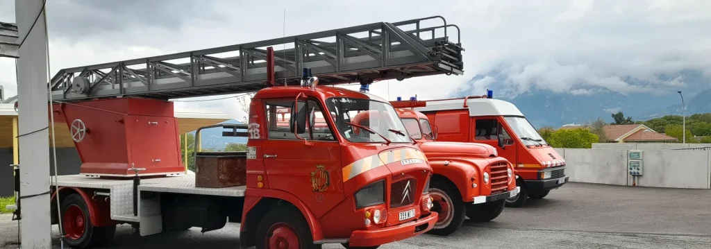 Trois camions de pompiers rouges sont stationnés devant une caserne, avec un paysage montagneux en arrière-plan sous un ciel nuageux. Un camion-échelle se distingue nettement.