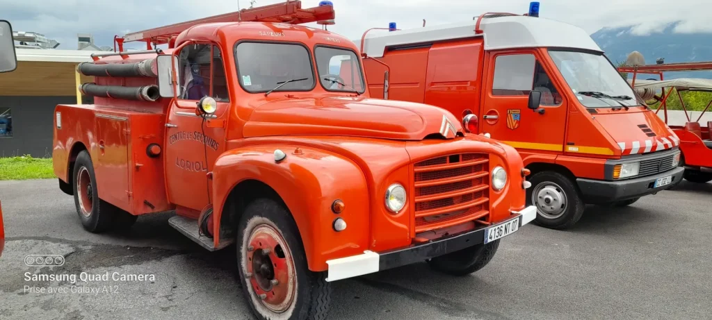 Un camion de pompiers rouge d'époque, avec ses tuyaux et portant l'inscription « Centre de Secours Loriol », est garé à côté d'un véhicule d'urgence moderne.