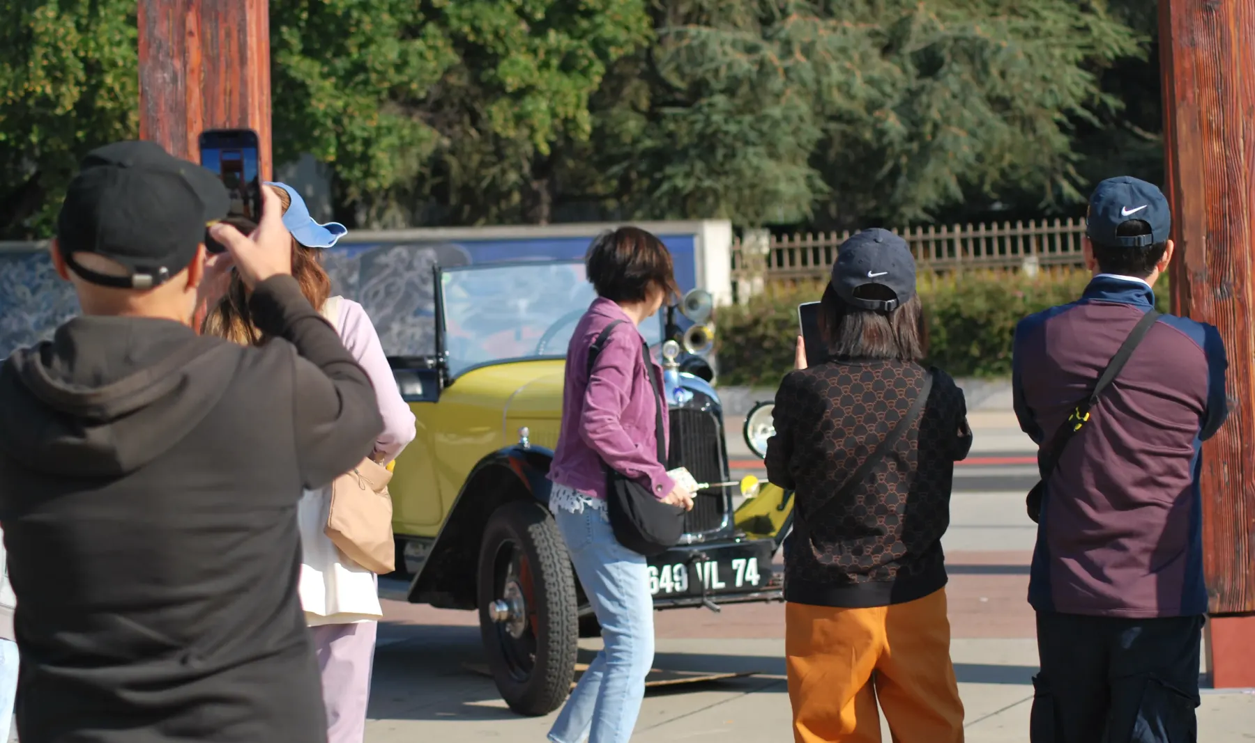 Un groupe de personnes en tenue décontractée prend des photos d'une voiture jaune vintage par une journée ensoleillée, entourée de verdure.