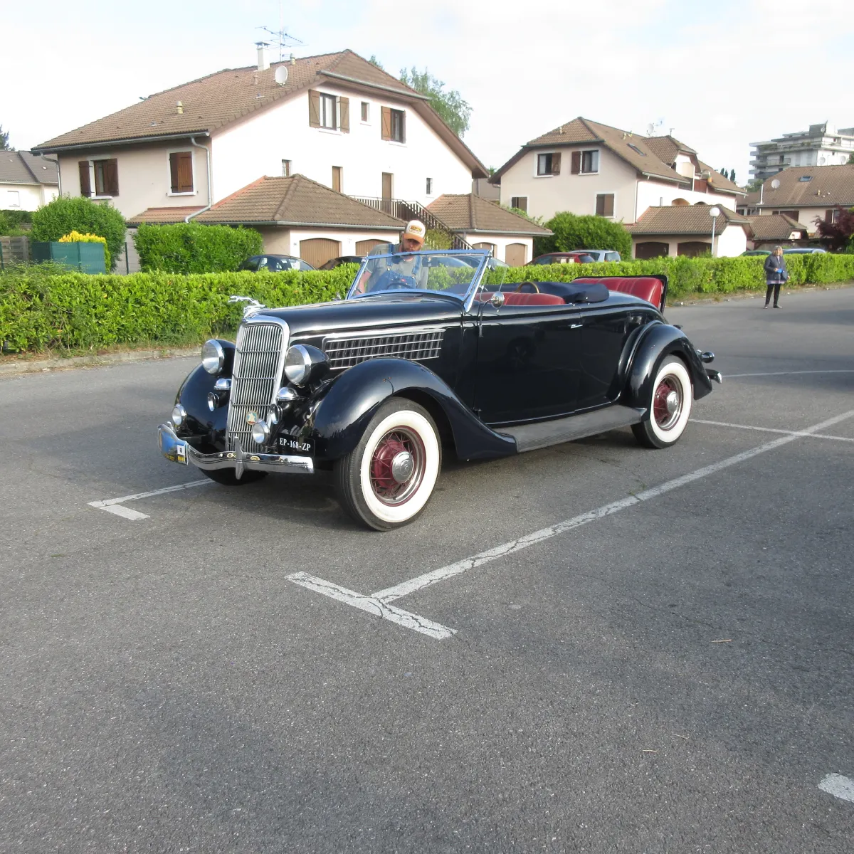 Une voiture décapotable classique des années 1930, avec une carrosserie noire, un intérieur rouge et des pneus à flancs blancs d'époque, est garée sur un parking, devant des immeubles résidentiels.